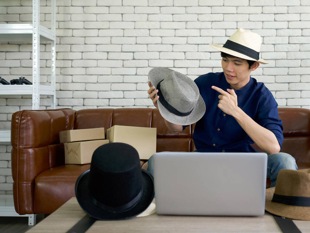 Joven asiático hombre vender varios estilos de ala sombrero en línea a través de la aplicación en el ordenador portátil. Concepto de pequeña empresa doméstica. - Foto, Imagen