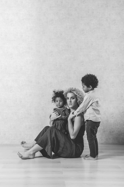 Caucasian young mother with curls hugs African American children. Happy mixed race family sitting on the wooden floor in the studio on gray background. Woman having fun with daughter, son, adoption - Foto, afbeelding