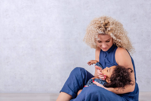 Caucasian young mother with curls hugs African American girl. Happy mixed race family sitting on the wooden floor in the studio on gray background. Woman holds in arms, caress daughter, adoption - Φωτογραφία, εικόνα