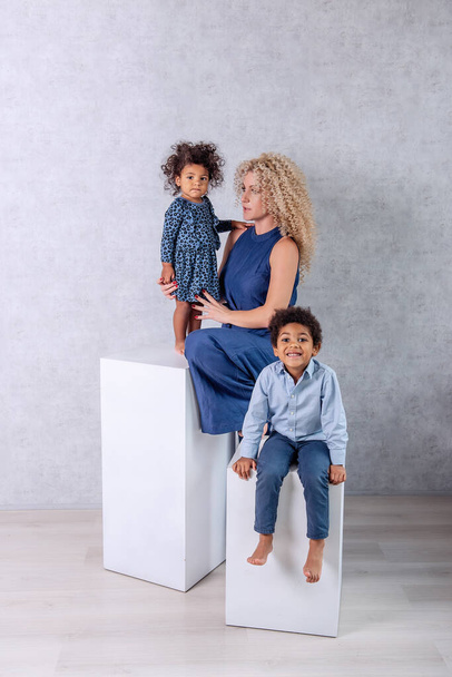 Caucasian young mother with African curls hugs African American children. Happy mixed race family sitting on white cubes in the studio on gray background. Woman holds daughter son in arms, adoption - Foto, Imagem