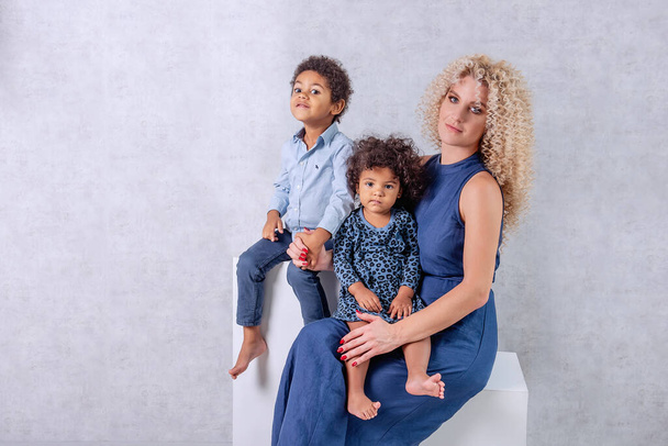 Caucasian young mother with African curls hugs African American children. Happy mixed race family sitting on white cubes in the studio on gray background. Woman holds daughter son in arms, adoption - Valokuva, kuva