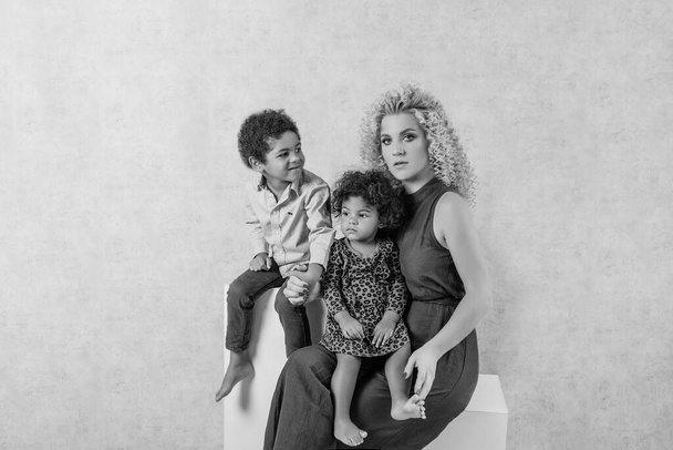 Caucasian young mother with African curls hugs African American children. Happy mixed race family sitting on white cubes in the studio on gray background. Woman holds daughter son in arms, adoption - Foto, Imagem