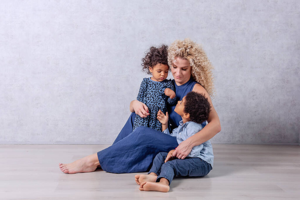 Caucasian young mother with curls hugs African American children. Happy mixed race family sitting on the wooden floor in the studio on gray background. Woman having fun with daughter, son, adoption - Photo, Image
