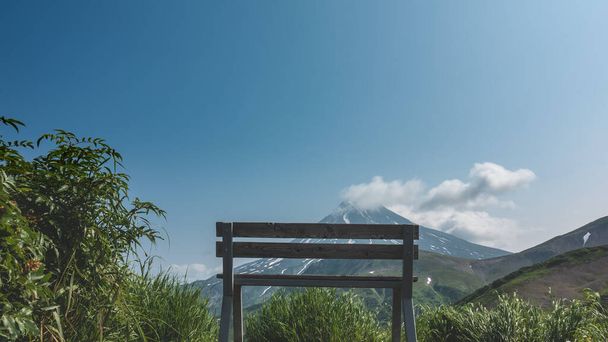緑の芝生の間に無塗装の木製のベンチがあります。先に、青い空、円錐火山に対して。斜面には雪が積もっている。上から雲だ。カムチャツカ。ヴィリュチンスキーのストラト火山 - 写真・画像