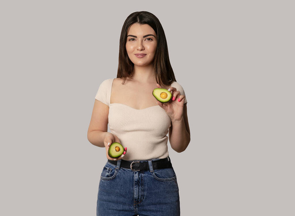 Smiling young woman holding a avocado, looking at camera, isolated grey background. healthy food. Horizontal view. Space for text. - Photo, Image