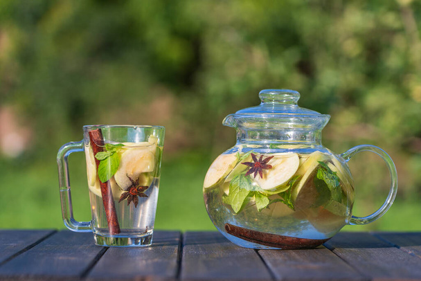 Healthy apple tea in a glass tea pot and mug in the summer garden on wooden table. Close up apple tea with cinnamon, cloves or mint leaves on nature background - Photo, Image