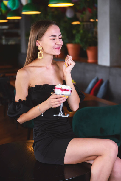 Beautiful sexy brunette woman in restaurant or cafe with ice cream tiramisu dessert. Fashion photo of young woman in black dress with hairstyle and makeup, eating dessert. Eat out concept. - Photo, image