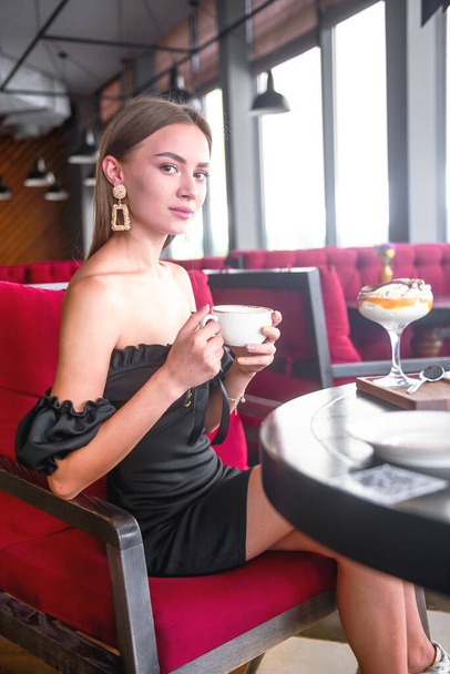 Beautiful sexy brunette woman in restaurant or cafe with ice cream tiramisu dessert and a cup of . Fashion photo of young woman in black dress with hairstyle and makeup eat out concept. - Фото, изображение