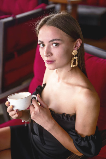 Beautiful sexy brunette woman in restaurant or cafe with ice cream tiramisu dessert and a cup of . Fashion photo of young woman in black dress with hairstyle and makeup eat out concept. - 写真・画像