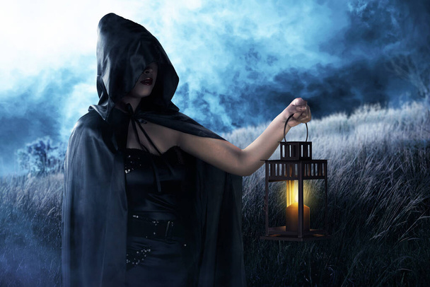 Asian witch woman with a black cloak holding a lantern standing on the field - Photo, image