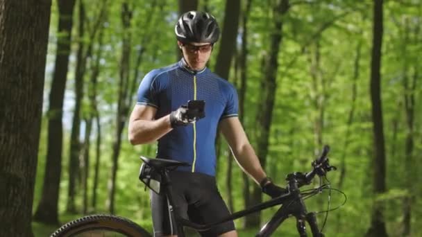 A man is looking through social networks on a smartphone. He is standing next to an MTB bike in the woods. Hes wearing cycling clothes. 4K - Footage, Video
