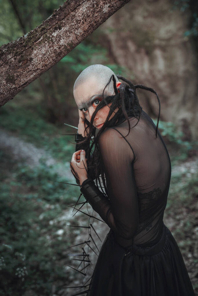 Mysterieuze spookachtige kale vrouw in een zwarte lange jurk. Gotische heidense dame in de buurt van de tempel en afgoden. De vampier wandelt in een diep donker mystiek bos.. - Foto, afbeelding