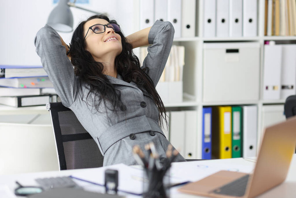 Счастливая довольная женщина отдыхает в офисе сидит с ноутбуком и держит руки за голову крупным планом - Фото, изображение