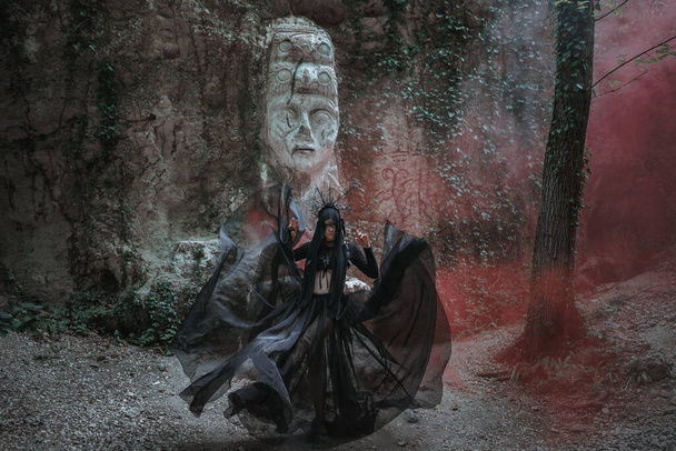 Jonge vrouw in heks staat in zwarte jurk en kroon op haar hoofd in donker en mistig bos. halloween kostuum voor donker feest. - Foto, afbeelding