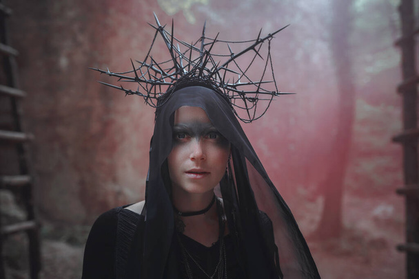 Jonge vrouw in heks staat in zwarte jurk en kroon op haar hoofd in donker en mistig bos. halloween kostuum voor donker feest. - Foto, afbeelding