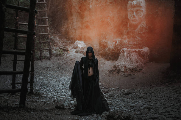 Eine junge Frau in Hexenkleidern steht in schwarzem Gewand im dunklen und nebligen Wald. Halloween-Kostüm für dunkle Party. - Foto, Bild