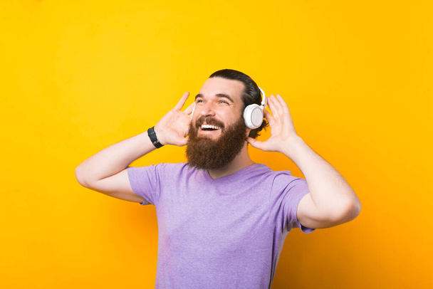 Foto di un uomo barbuto felice che sorride ascoltando musica in cuffia, sfondo giallo della parete - Foto, immagini