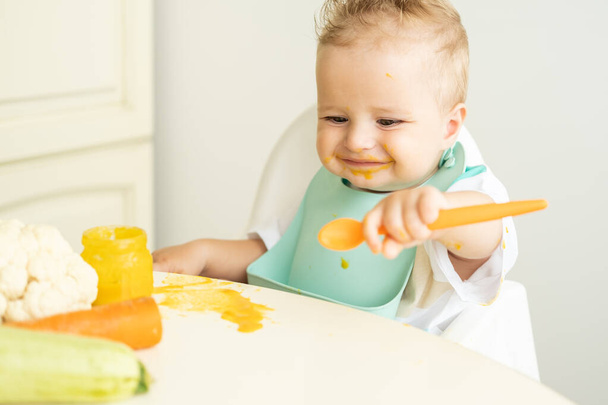 funny baby boy in bib eating vegetable puree with spoon sitting in child chair. Child learn to eat by himself. - Фото, зображення