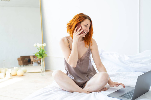 atractiva mujer jengibre de mediana edad en pijama sentado en la cama utilizando el teléfono y el ordenador portátil. - Foto, imagen