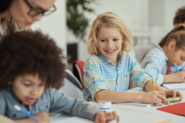 Aktywny mały uczeń uśmiechający się i patrzący na swojego kolegę z klasy siedzącego przy stole w klasie podstawowej razem z innymi dziećmi - Zdjęcie, obraz
