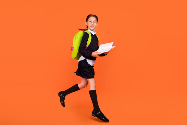 Full body photo of cheerful brunette braids haired girl jump up hold book wear bag isolated on orange color background - Фото, изображение