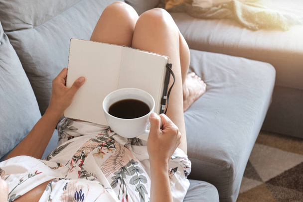 Женщина лежит на диване с кофейной чашкой и смотрит в блокнот, место для текста - Фото, изображение