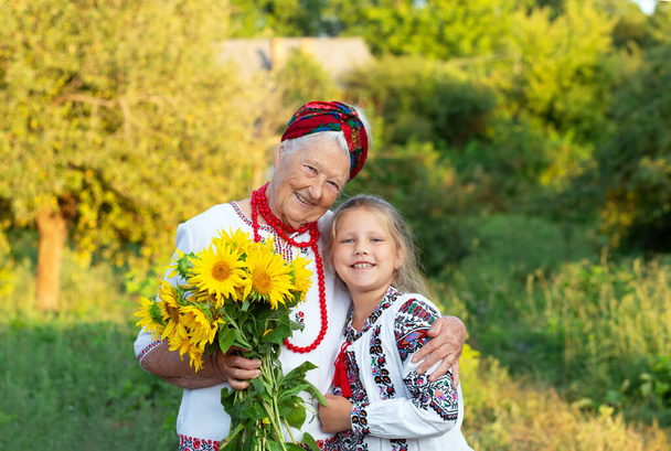 Gri saçlı büyükanne ve kız torun. İkisi de işlemeli tişörtler giyip bir buket ayçiçeğiyle mutlu bir şekilde gülümsüyor. Bağımsızlık Günü, Ukrayna Anayasası - Fotoğraf, Görsel