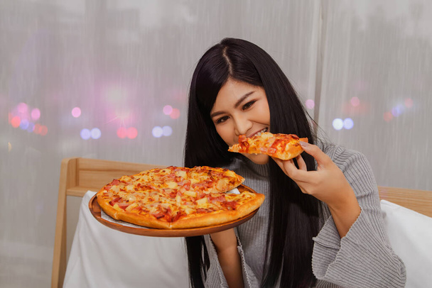 Schöne Asiatin isst Pizza Völlerei ist Abendessen, vor dem Schlafengehen gegessen, Essen im Bett in ihrem Haus mit Vergnügen: Frau bestellt während der Covit-19-Epidemie online Pizza für die Lieferung nach Hause - Foto, Bild