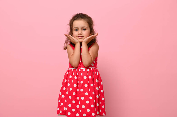Adorable niña en vestido rosa con lunares posa en la cámara, de pie sobre un fondo rosa y sosteniendo su barbilla con las manos. Retrato de una hermosa chica con emociones alegres positivas - Foto, imagen