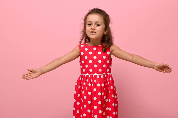 Улыбающаяся счастливая маленькая очаровательная девочка с протянутыми руками в розовом платье с горошек, смотрящая в камеру, позируя на розовом фоне. Портрет добродушного радостного, приветливого, дружелюбного ребенка - Фото, изображение