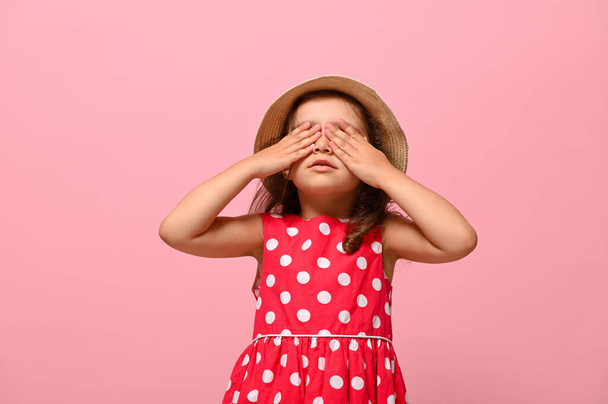 Confident portrait of gorgeous baby girl dressed in summer polka dots dress and straw hat , covering her eyes with her hands, posing over pink background with space for text for advertising - Foto, afbeelding