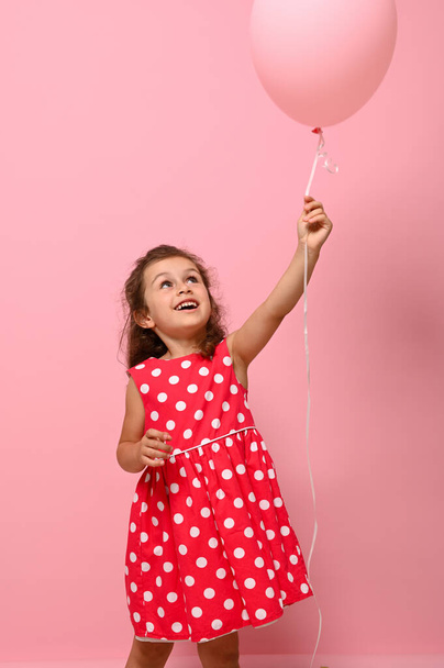 Urocza piękna 4-letnia śliczna solenizantka w różowej sukience w kropki raduje się patrząc na różowy balon w podniesionej ręce, odizolowany na różowym tle z przestrzenią do kopiowania. Prawdziwe emocje z dzieciństwa. Pojęcia - Zdjęcie, obraz