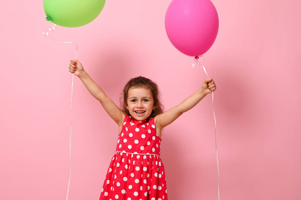 Birthday Baby girl dressed in pink dress with polka dots pattern raising her arms up with colorful balloons in her hands, smiling looking at the camera, isolated on pink background with copy space - Fotó, kép