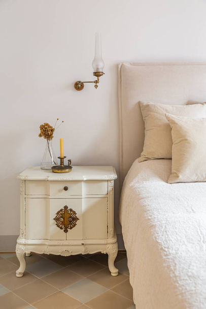 Interieur van gezellig huis in retro stijl. Klassieke slaapkamer met kaars en bloemen op houten nachtkastje in de buurt van comfortabel bed. - Foto, afbeelding