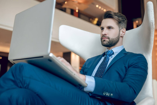 Takım elbiseli bir adam koltukta oturuyor ve dizüstü bilgisayarla çalışıyor. - Fotoğraf, Görsel