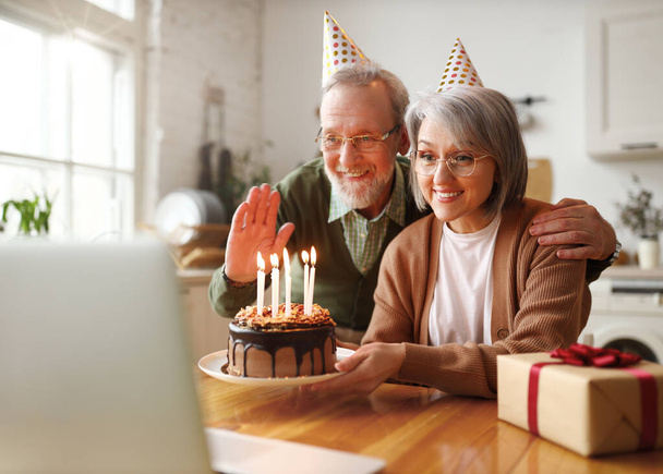 Krásný starší pár ve straně klobouky slaví narozeniny doma on-line na notebooku, dostávají gratulace od rodiny koryto video chat, šťastný muž mává na kameru, žena drží čokoládový dort - Fotografie, Obrázek