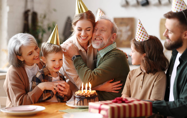 Χαρούμενος ευτυχισμένος ηλικιωμένος που αγκαλιάζεται με τη μικρή του κόρη και αισθάνεται ευγνώμων ενώ γιορτάζει τα γενέθλιά του με μεγάλη αγαπημένη οικογένεια, κάθεται μαζί με καπέλα κόμμα στο τραπέζι με κέικ και κουτί δώρου - Φωτογραφία, εικόνα