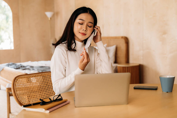 Young teenage Asian woman wears sweater using computer laptop working from home indoors online. Attractive girl removing a face mask. - Valokuva, kuva
