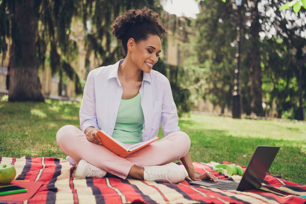 Full length photo of young african woman happy positive smile use laptop lecture college write copybook study online outdoors - Foto, afbeelding