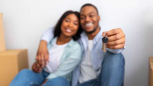 Новый дом. Возбужденные чернокожие супруги показывают ключи, сидя в собственном доме после переезда, сосредоточиться на ключе, панорама - Фото, изображение