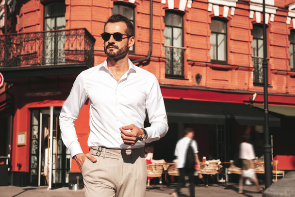 Portrait of handsome confident stylish hipster lambersexual model.Modern man dressed in white shirt and trousers. Fashion male posing in the street background in Europe city at sunset. In sunglasses - Foto, afbeelding
