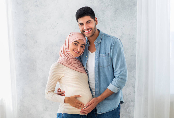 若いです妊娠中のアラブ配偶者の肖像,抱擁と笑顔でカメラ,彼の妻腹を抱いて愛するイスラム教徒の男 - 写真・画像