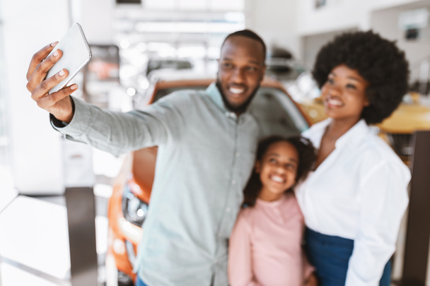 Joyful black family taking smartphone selfie near their new luxury car at auto dealership, selective focus - Photo, Image