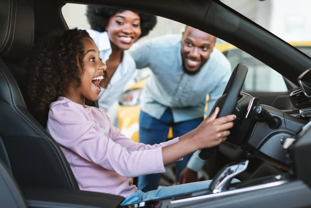 Ragazza nera seduta al sedile del conducente della nuova auto, urlando di eccitazione, aiutando i suoi genitori a comprare il veicolo presso la concessionaria auto - Foto, immagini