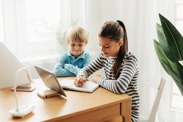Küçük erkek kardeşiyle aynı masada oturan bir kız öğrenci bir tablet üzerinde ödev yapar ve bir deftere not alır. - Fotoğraf, Görsel