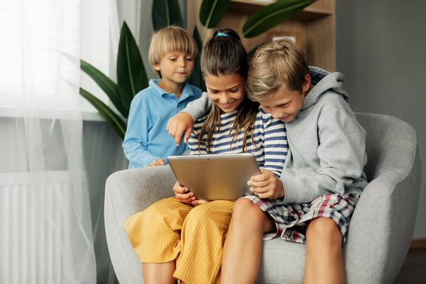 The beautiful dark-haired sister and her brothers are sitting at home on the couch, playing games on the tablet and chatting with friends via video. Modern technology and spending time at home - Zdjęcie, obraz