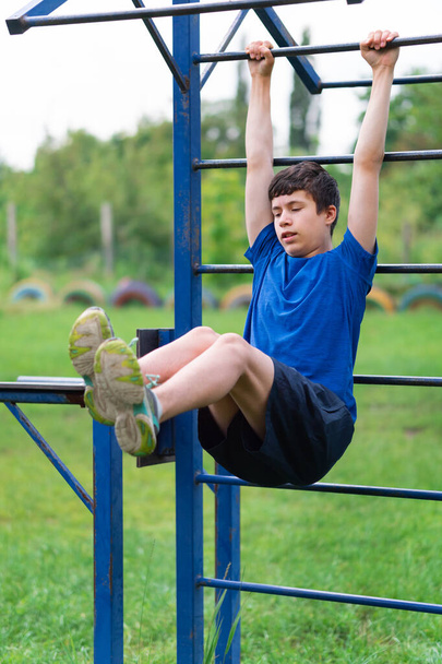teenage boy exercising outdoors, sports ground in the yard, he hangs on the horizontal bar, raises his legs and pumps his abs, healthy lifestyle - Photo, Image