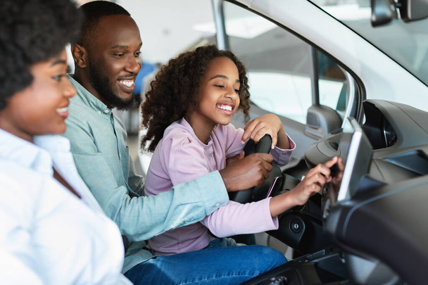 Vrolijke zwarte familie controleren auto salon voor het kopen van nieuwe auto, zitten in de auto, gaan op proefrit - Foto, afbeelding
