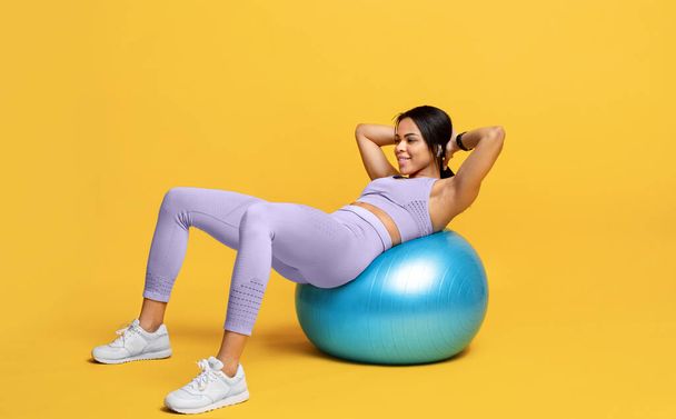 Sportliche schwarze Frau macht Bauchmuskelübungen auf Fitnessball vor gelbem Studiohintergrund, freier Raum - Foto, Bild