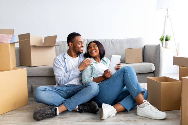 Milující černošky rodina pomocí digitálního tabletu a objímání při sezení na podlaze v novém domě mezi unpacked boxy - Fotografie, Obrázek